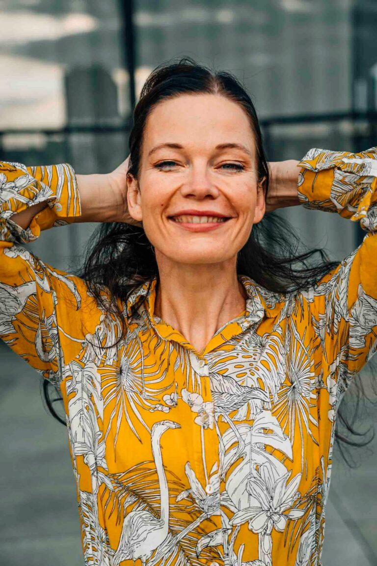 Dark-haired woman smiling with her hands behind her head outdoors