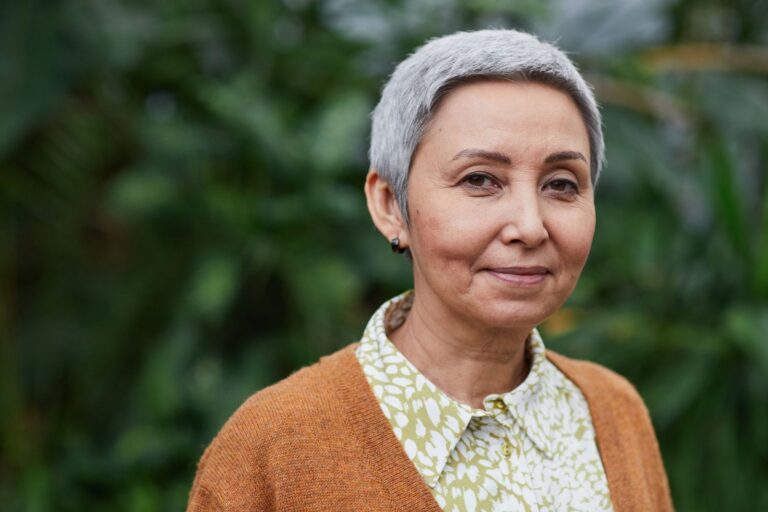 A woman in her sixties with short grey hair, outdoors among green leaves