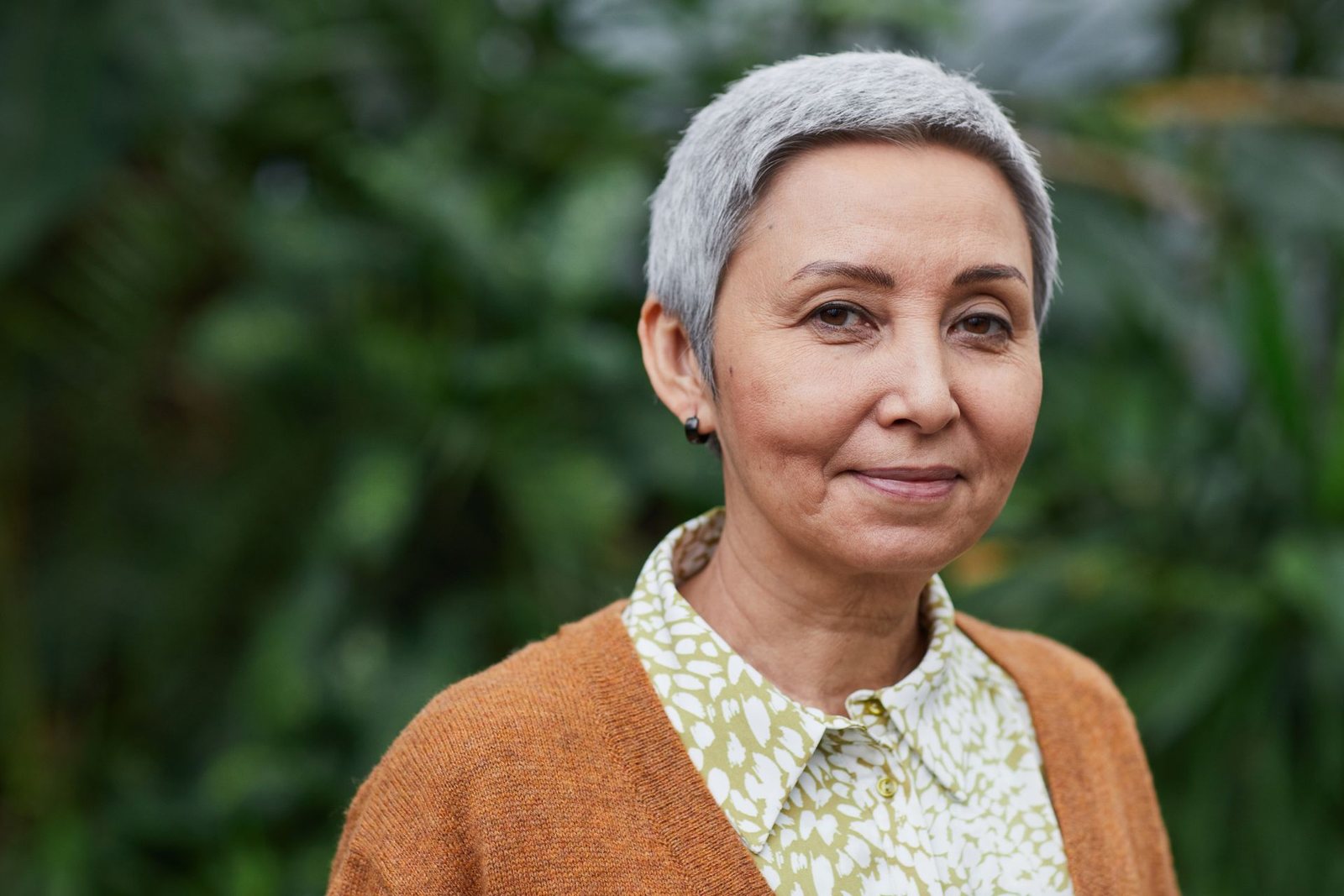 A woman in her sixties with short grey hair, outdoors among green leaves