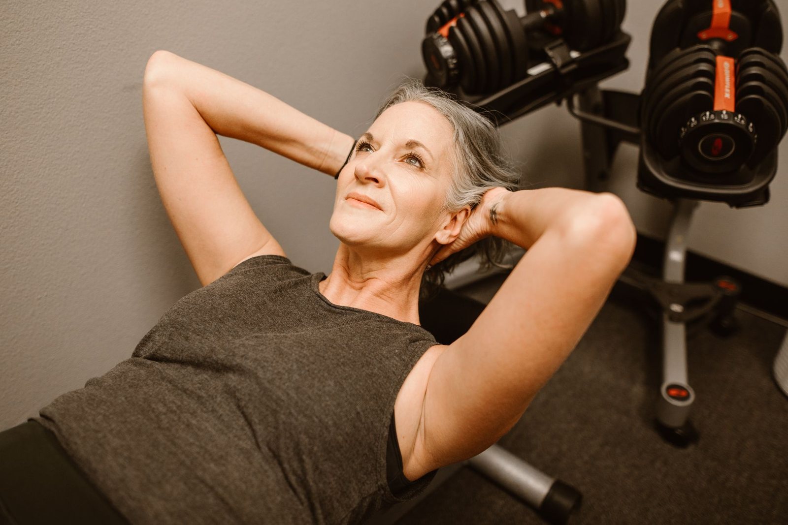 A woman in her fifties with grey hair doing a sit-up on a bench beside dumbbells