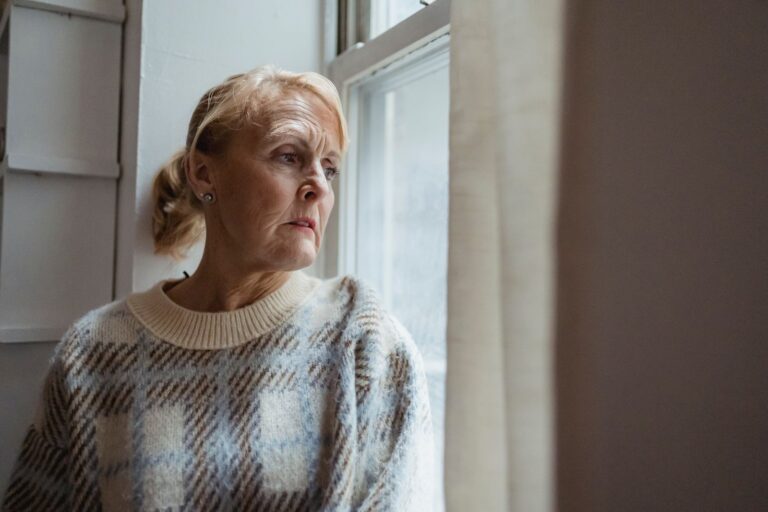 A woman in her sixties in a knitted jumper looking out of a window