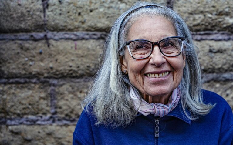 A woman of about seventy with long grey hair and glasses, smiling outdoors