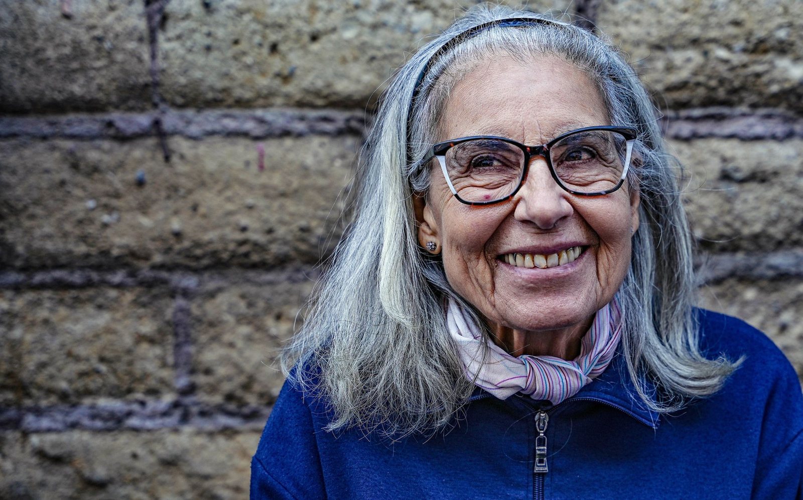 A woman of about seventy with long grey hair and glasses, smiling outdoors